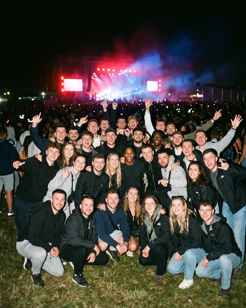 The same man standing small in the middle rows of a large group photo taken at a night-time outdoor concert.