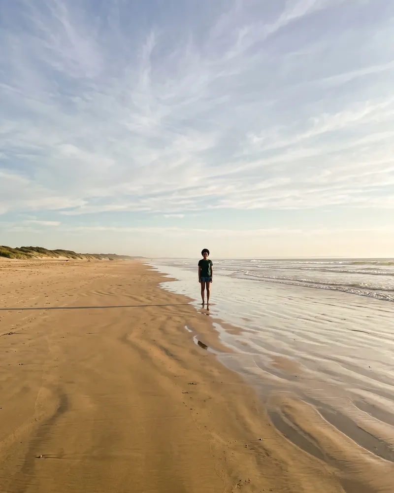 The same woman standing far away at the water’s edge on a wide beach, tiny in the frame under a big sky.