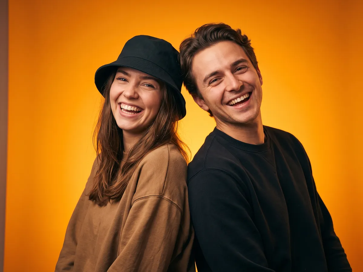 A smiling young woman in a black bucket hat and a grinning young man pose back-to-back against a bright orange studio backdrop.