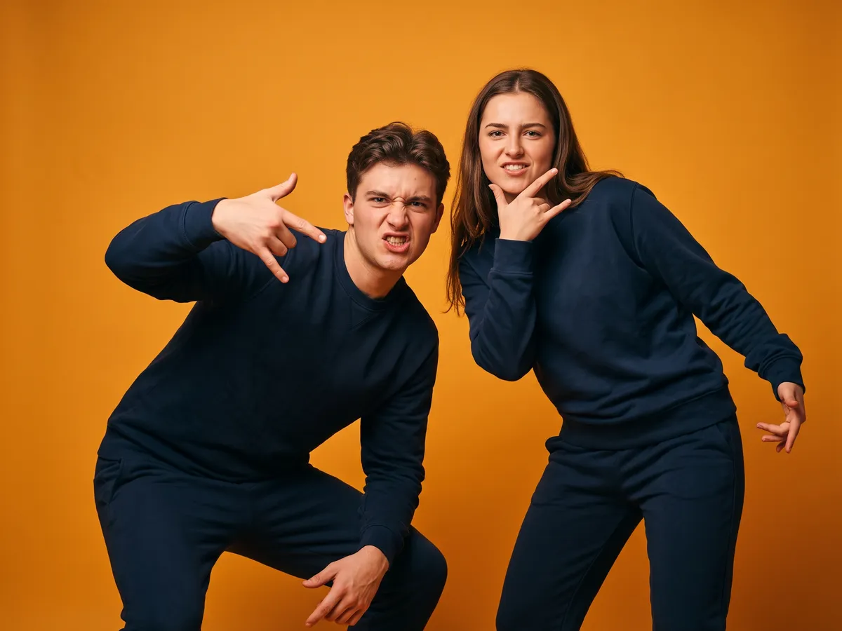 A young man and a young woman in matching navy tracksuits make exaggerated rap hand-sign poses against an amber-orange studio wall.
